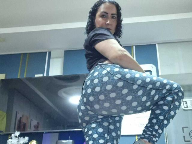 SilverCool69 with curly black hair, wearing a black shirt and blue polka dot pants, standing with one leg raised, showing her big ass and looking back.