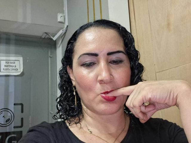 SilverCool69, con i capelli neri ricci, rossetto rosso e trucco occhi scuro, fa la smorfia con un dito sulla labbra inferiore, indossando una maglietta nera e orecchini dorati, in un selfie..