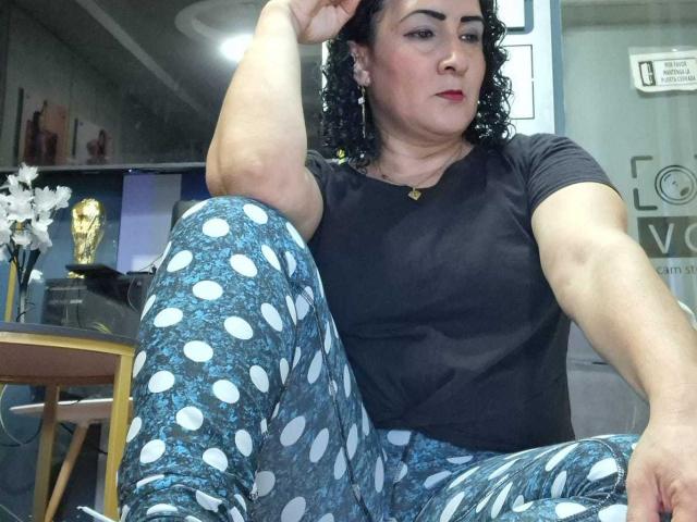 SilverCool69, com cabelo preto encaracolado, batom vermelho, camiseta preta e pijama azul com bolinhas, sentada com um braço levantado, mostrando um braço musculoso e uma expressão confiante..