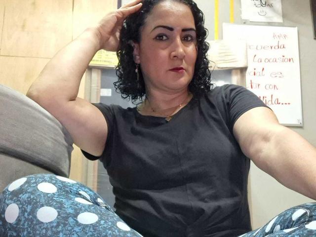 SilverCool69 ist eine kurvige Frau mit schwarzen, lockigen Haaren, die ein schwarzes Hemd trägt und mit einem Arm erhoben sitzt. Sie blickt in die Kamera und hat einen ernsten Gesichtsausdruck..