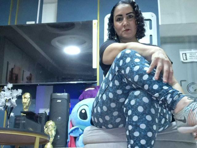 SilverCool69 sits on a couch in polka dot pajamas, black shirt, short curly hair, tattoo on ankle, arm resting on knee, TV and stuffed toys in background.
