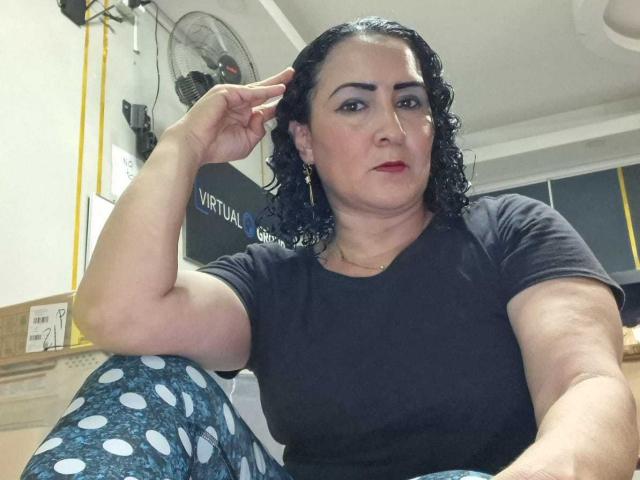 SilverCool69 ha i capelli neri ricci, il rossetto rosso e indossa una maglietta nera con pantaloni blu a pois, seduta con una mano tra i capelli, mentre guarda la fotocamera..