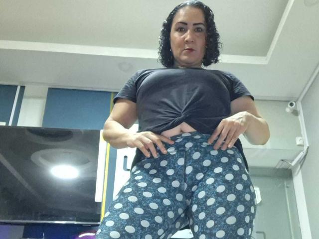 SilverCool69 with curly black hair, wearing a tied black shirt and blue polka dot pants, standing with hands on hips, showing a flat stomach and small tits.