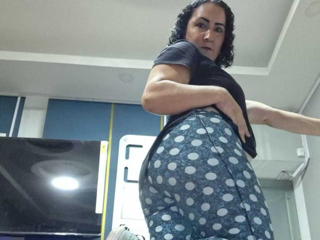 SilverCool69 with curly black hair, wearing a black shirt and blue polka-dot pants, showing her ass to the camera in a low-angle shot.