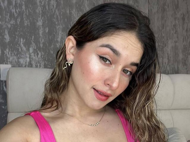 MilaGonz mit feuchtem, braunem Haar, einem pinken Oberteil, einer kleinen Nase, heller Haut, grünen Augen, dezentem Make-up, Ohrringen und einer Halskette, mit einem leichten Grinsen, die in die Kamera schaut, in einem Selfie..