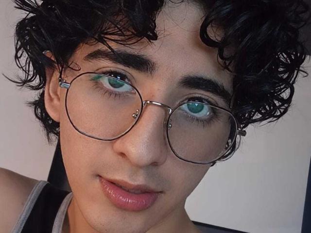 ElliotNoir has curly black hair, green eyes, and wears round glasses with metal earpieces. Their pink lips are slightly parted. They have a light skin tone and a subtle nose piercing.
