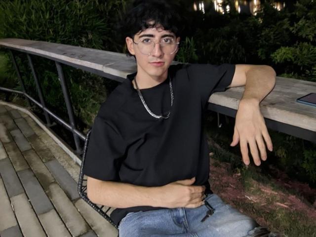 ElliotNoir with curly black hair, wearing glasses, a black t-shirt, and blue jeans, sits on a wooden bench at night, hand on belly, chain necklace, relaxed, looking at camera.