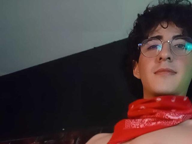ElliotNoir with curly black hair, glasses, and a red bandana, smiling at the camera, showing bare shoulders. A light green reflection is on ElliotNoir's glasses.
