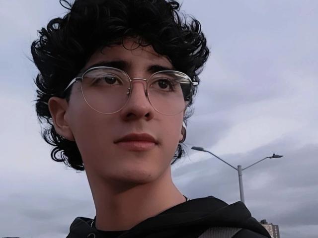ElliotNoir with curly black hair, round glasses, and a black hoodie, showing a calm face with slight pink lips, under a cloudy sky.