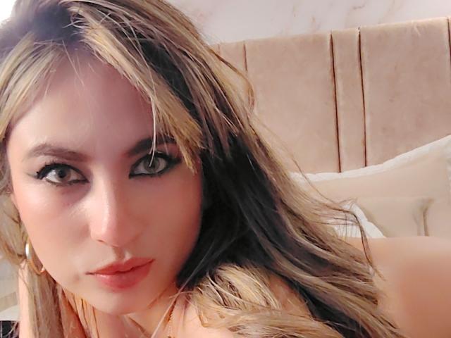 AmiiRosse has long, wavy brown hair, full lips with red lipstick, and dark eyeliner, staring into the camera with green eyes, wearing gold earrings, in a bedroom.