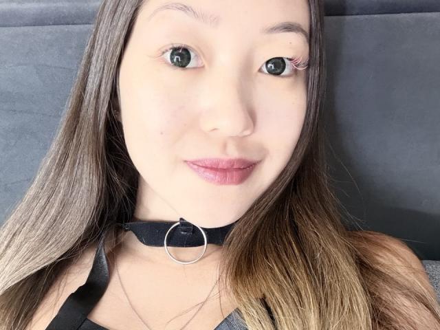RenyLime has light skin, green eyes, and brown hair, wearing a black choker with a silver ring, and pink lipstick, sitting with a slight smile. Boobs not visible.