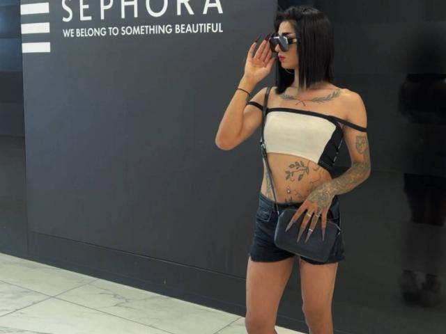 Iris, avec des tatouages, une coupe de cheveux courte noire, portant un haut blanc à une épaule, un short en jean, un sac bandoulière noir, des lunettes de soleil, tenant un téléphone, debout, fond noir, sol en marbre, tenant un téléphone, regardant la caméra..