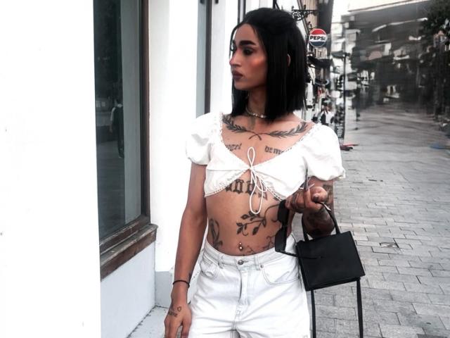 IrisIrs with black hair, white crop top showing her tits and tattoos, white pants, black bag, standing outside, looking to the side, street in background.