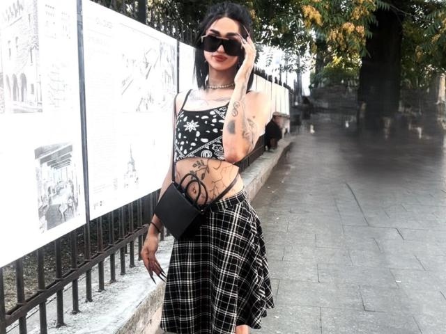 Iris, con tatuaggi, occhiali da sole neri, rossetto rosso, top corto bianco e nero, gonna a quadri nera, che tiene una pochette nera, in piedi su un marciapiede bagnato, appoggiata a una recinzione di metallo, con un albero sullo sfondo..