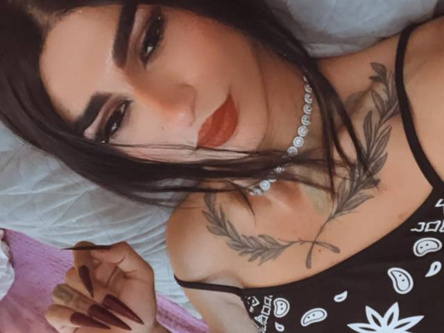 Iris, avec ses cheveux foncés, ses sourcils audacieux, son rouge à lèvres rouge, son haut noir, son collier et son tatouage floral sur la poitrine, se tient les cheveux. Ses seins sont visibles, mais ses fesses ne le sont pas..