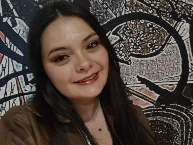 Katherin, con cabello negro lacio, ojos marrones y un pequeño aro en la nariz, sonriendo con labios brillantes, vistiendo una blusa marrón y un collar rojo diminuto..