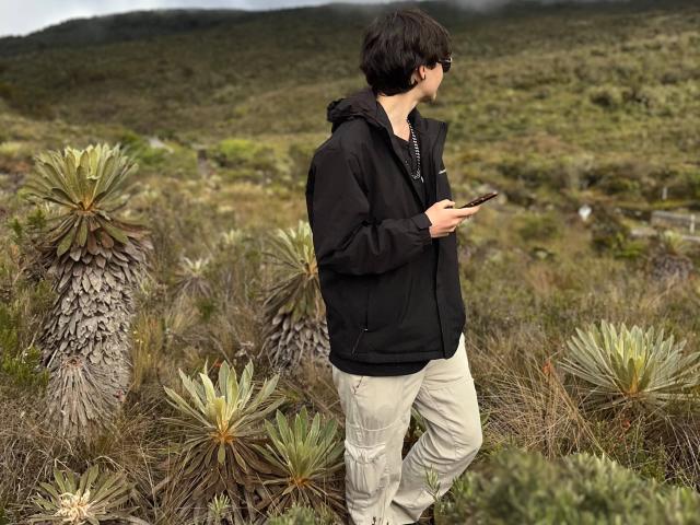 LissyLuxe stands in a wild, green, rocky landscape holding a smartphone, wearing a black hoodie and beige cargo pants, with short brown hair and dark sunglasses.