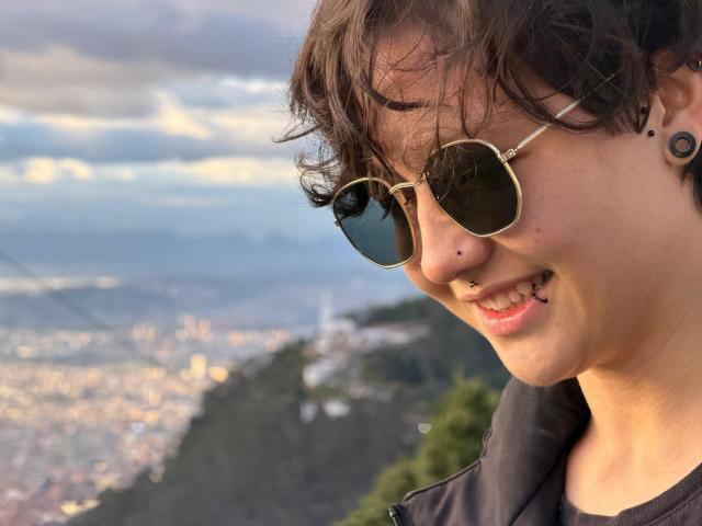 LissyLuxe has short, curly brown hair, wears round sunglasses, and a nose piercing, smiling with a lip ring, showing black ear plugs, in a sunny, mountainous, cityscape background.