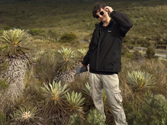 LissyLuxe stands in a desert with green cactus, wearing a black jacket, beige pants, and sunglasses, holding a phone, with curly brown hair, and a slightly annoyed look.