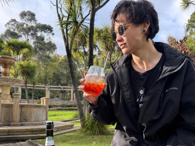 LissyLuxe with short black hair, sunglasses, black jacket, and black shirt, holding a glass of orange liquid, standing next to a bottle, in a sunny garden.