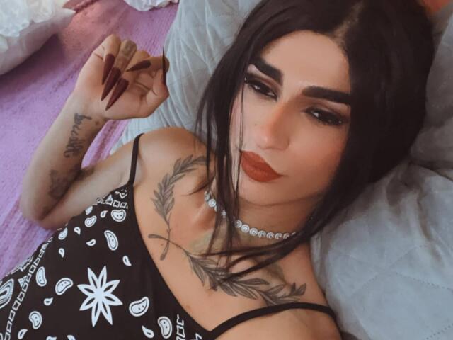 IrisIrs has dark hair, thick eyebrows, red lipstick, tattoos on her chest and arm, and is wearing a black, white-patterned top with spaghetti straps, lying on a blue pillow.
