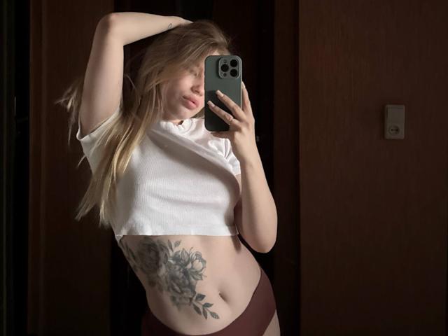 MariluChouinard with blonde hair, white crop top, and maroon panties, showing a large floral tattoo on her right side, taking a mirror selfie with her phone.