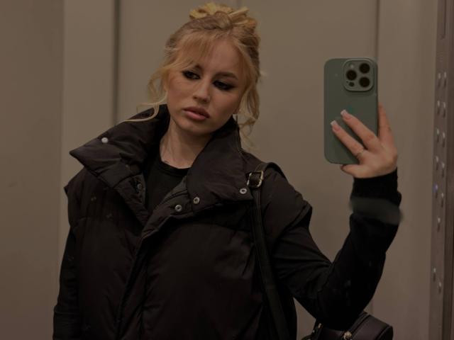 MariluChouinard taking a mirror selfie with a green iPhone, wearing a black puffy jacket, blonde hair in a messy bun, holding a black bag, looking slightly annoyed.