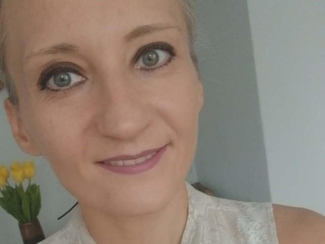 ViolettaLoves ha gli occhi verde chiaro, un leggero sorriso e rossetto rosa. Indossa una camicetta di pizzo bianco, con dei tulipani gialli sullo sfondo. Il suo viso è di carnagione chiara..