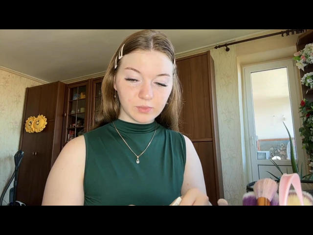 ConchitaMosburg with light brown hair, green sleeveless top, and gold necklace, looking down, holding a white object in a room with wooden cabinets and a yellow wall decoration.