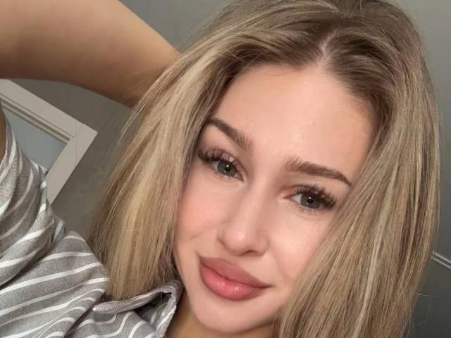 MilagrosEdmisten has blonde hair, green eyes, and full pink lips. She's wearing a striped shirt, smiling with a slight blush, and has long, thick eyelashes.