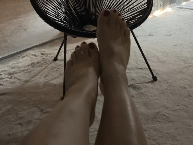 Dreia's bare feet with red-painted toenails are crossed in front of a black, circular, wire-frame chair on a sandy floor.