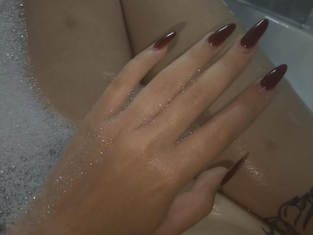 Dreia's hand with long, red, glossy nails rests on her wet, soapy thigh in a bathtub. Her tattoo is partially visible on her side.