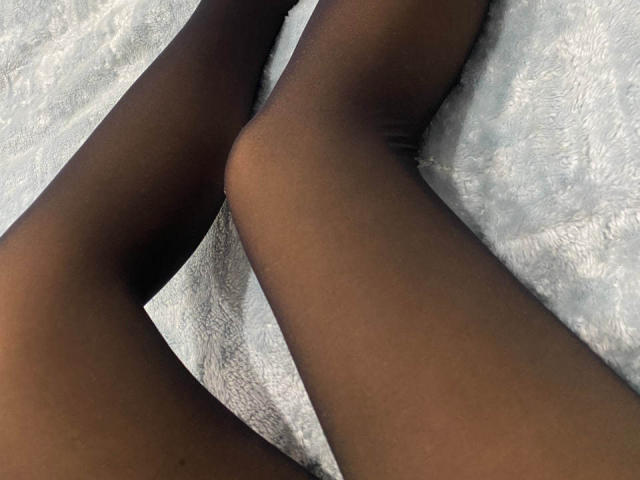 JessikaBernskoetter's dark brown, sheer pantyhose-covered legs are crossed at the ankles on a white, textured fabric. The focus is on her smooth, toned thighs.