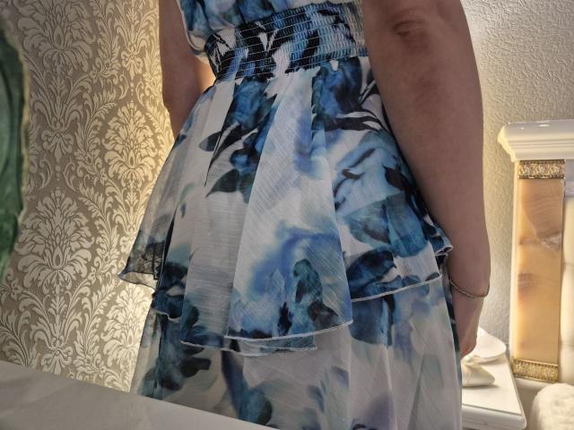 Anshante's blue and white floral dress has a flared, multi-layered skirt with a visible waistband. Her arm is slightly raised, showing a light skin tone.
