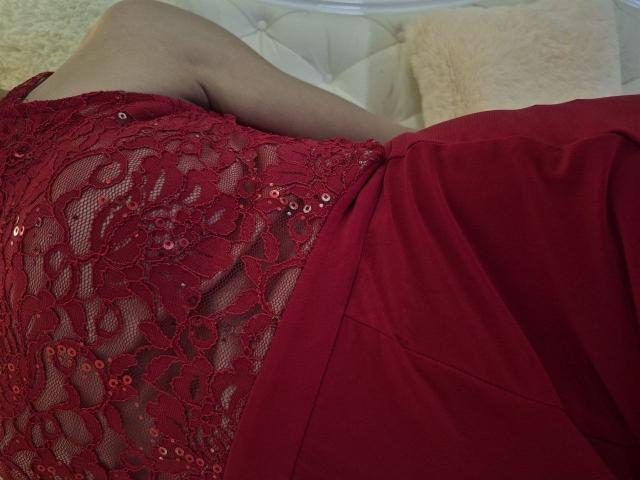 Anshante's rode kanten jurk met pailletten, waarin haar blote borsten zichtbaar zijn, ligt op een wit bed met knopen. Haar arm is zichtbaar, de jurk bedekt gedeeltelijk haar rode ondergoed..