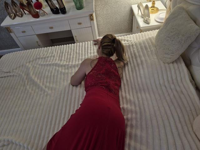 Anshante ligt op haar buik op een bed, ze draagt een rode kanten jurk, haar billen zijn omhoog, haar armen zijn vooruit, haar blonde haar zit in een paardenstaart, het bed heeft witte ribbenstructuur, en er staat een witte nachtkastje met gouden items..