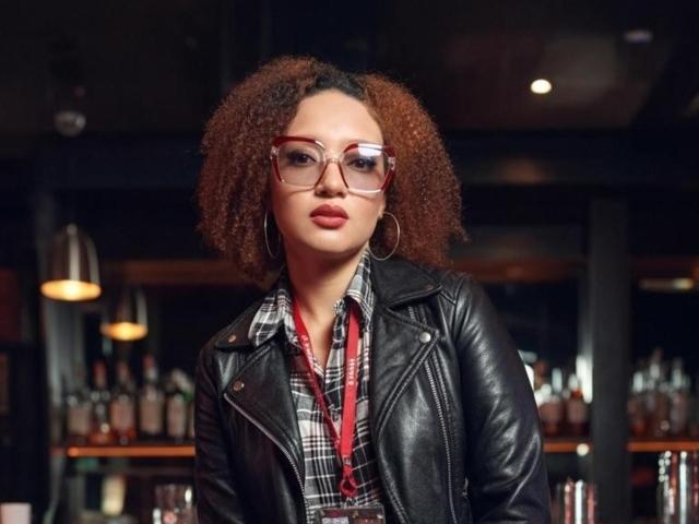 VioletaSwitch with curly brown hair, red glasses, and hoop earrings, wearing a black leather jacket and plaid shirt, standing in a bar, looking straight at the camera.