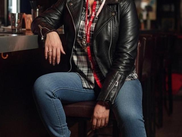 VioletaSwitch with black leather jacket, plaid shirt, and blue jeans, sitting on a bar stool, hand on knee, red lanyard, bar counter in background, face out of frame.