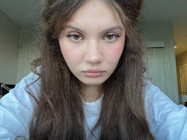 TanaFietek heeft lang, rommelig bruin haar, bleke huid en eyeliner met een vleugvorm. Ze draagt een witte blouse, heeft roze wangen en een neutrale, lichtjes fronsende uitdrukking..