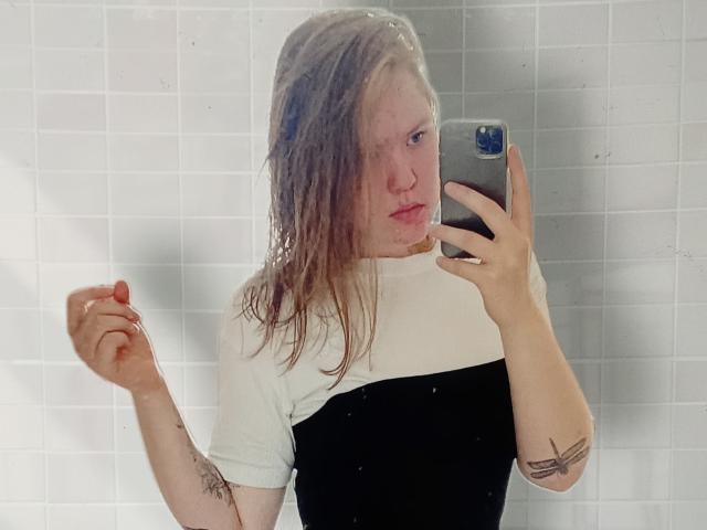 VioletteRo avec des cheveux blonds clairs, un haut blanc et noir, tenant un téléphone, un selfie pris devant un miroir, bras droit levé, tatouage sur le bras gauche, expression sérieuse, fond avec un mur en carreaux blancs..