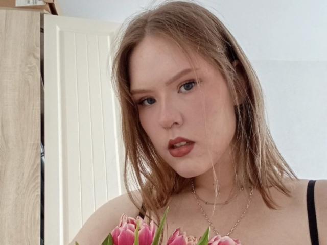 VioletteRo with light brown hair, blue eyes, and pink lipstick, holding pink tulips, wearing a black top and silver necklace, looking slightly tired.