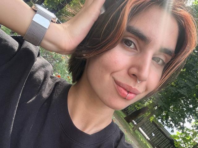 TameikaMacclellan with short brown hair, nose ring, and lip piercing, wearing a black shirt, touching her head, sunbeam, and gray watch on her wrist.