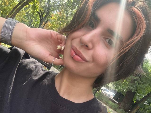 TameikaMacclellan with short brown hair, nose ring, lip ring, black shirt, sunlight, and green trees in background, smiling, hand touching ear, wristband visible.