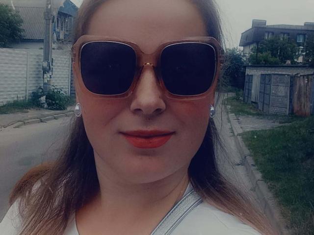 MelodyAmber, met bruine aviatorzonnebril, rode lippenstift en een wit shirt, draagt oorbellen, heeft bruin haar en een licht mollig gezicht, en staat buiten op een straat in een buitenwijk..