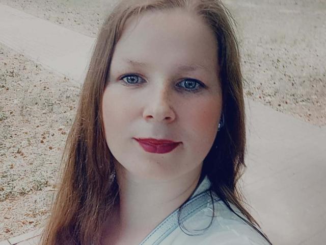 MelodyAmber tiene ojos azules, lápiz labial rojo y cabello largo castaño, lleva una camisa azul claro, con pequeños aretes, y mira directamente a la cámara con una ligera sonrisa..