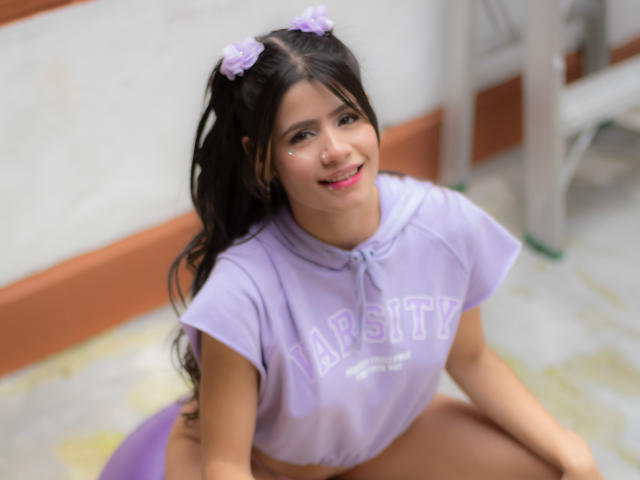 SheylaSpooner with purple puffy shirt, lilac hair bobbles, nose glitter, smiling, squatting, visible purple panties, long black hair, cute, blurred background, light skin, pink lipstick.