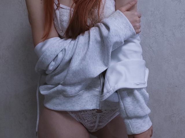 SheritaChieng with red hair, white lace panties, and a gray hoodie pulled down, exposing her tits and pussy. Her hand covers her chest, showing a modest, sexy pose.