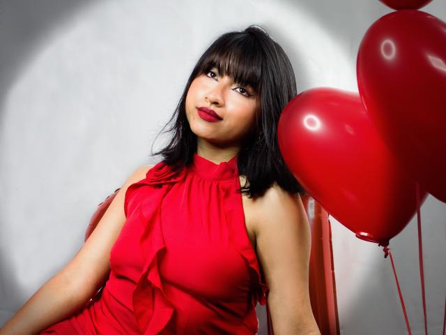 DariaHotst with black hair, red lipstick, and red dress, sitting on chair with red heart balloons, looking slightly up, has medium tits, cute, and sexy.