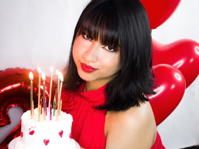 DariaHotst with black bob, red lipstick, and red dress, holding a white cake with candles, red heart balloons in the background, smiling at the camera.