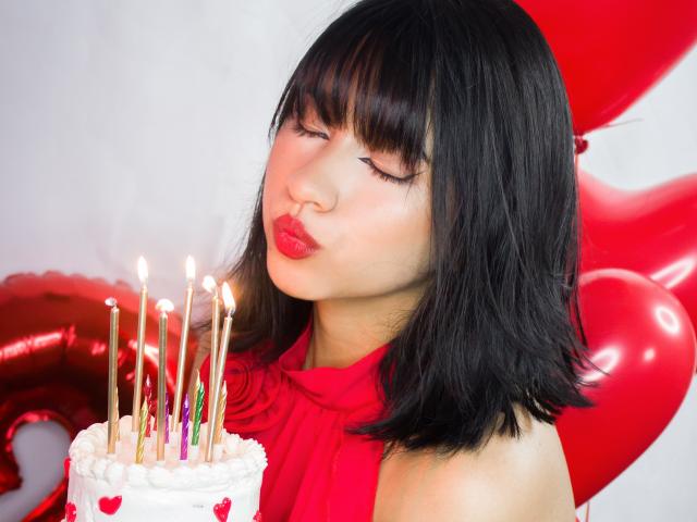 DariaHotst with black hair, red lipstick, and closed eyes, blowing kiss on white cake with candles, wearing red halter, red heart balloons in background.